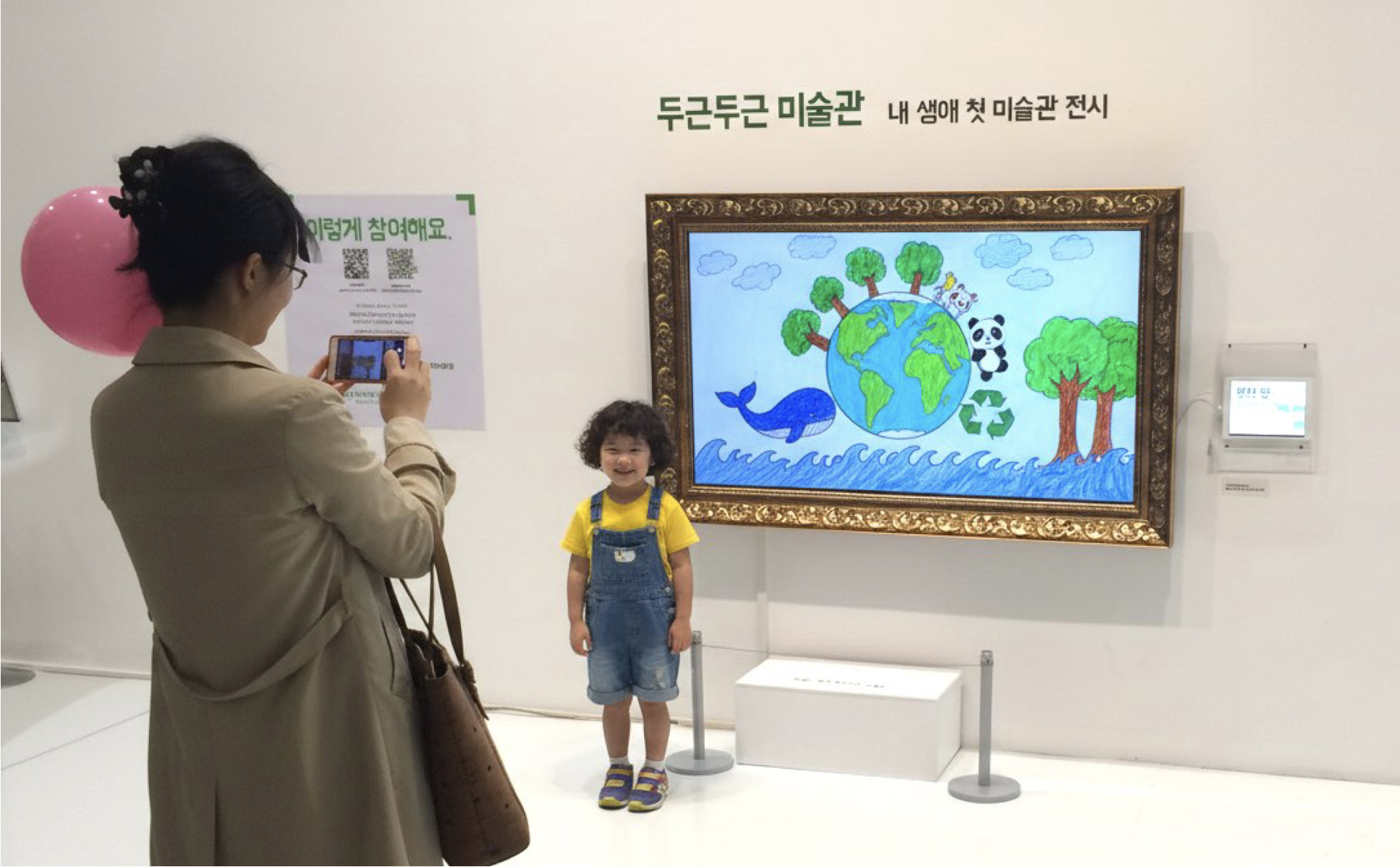 A child smiling in front of their artwork displayed on a digital frame at a dgdgM exhibition